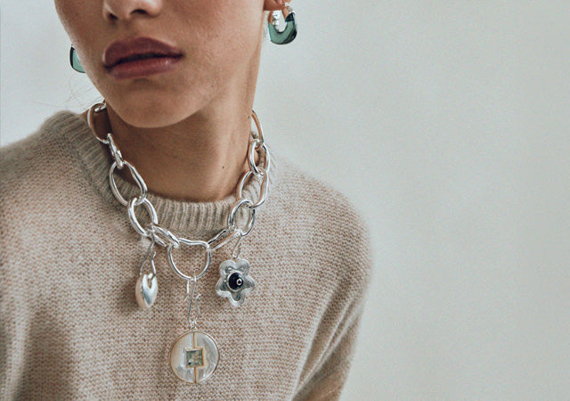 Model on grey backdrop wears tan sweater with silver necklace and charms