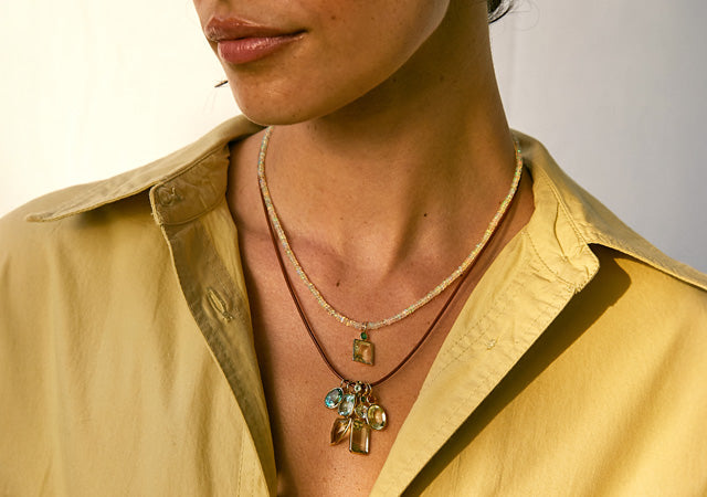 Model wears tan shirt with tiny opal and leather cord necklaces and charms