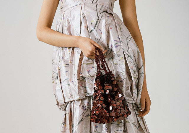 Model on grey backdrop wears lavender dress and holds Gala Bag in Garnet