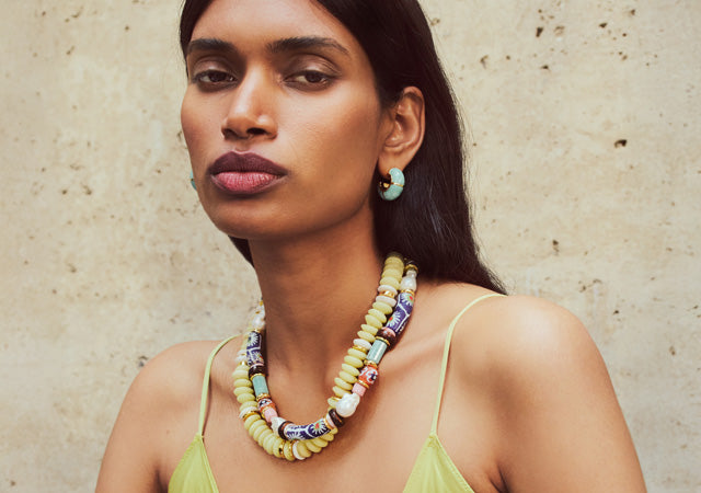 Model on tan stone backdrop wears Souvenir and Laguna Necklaces with Zellige Hoops