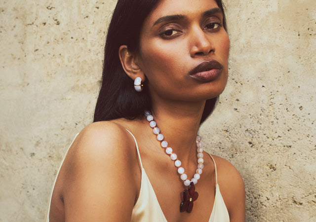 Model on stone backdrop wears strappy tank with Vinca Flower Necklace and Zellige Hoops