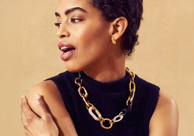 Model on tan backdrop wears black sleeveless sweater with chain link necklace
