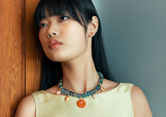 Model on wood panel and grey backdrop wears pale lime top with blue charm necklace and stud earrings
