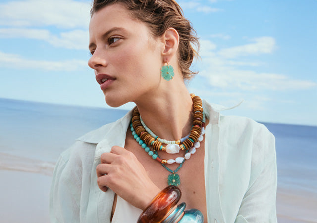 Model on beach wears stack of beaded necklaces and resin bracelets
