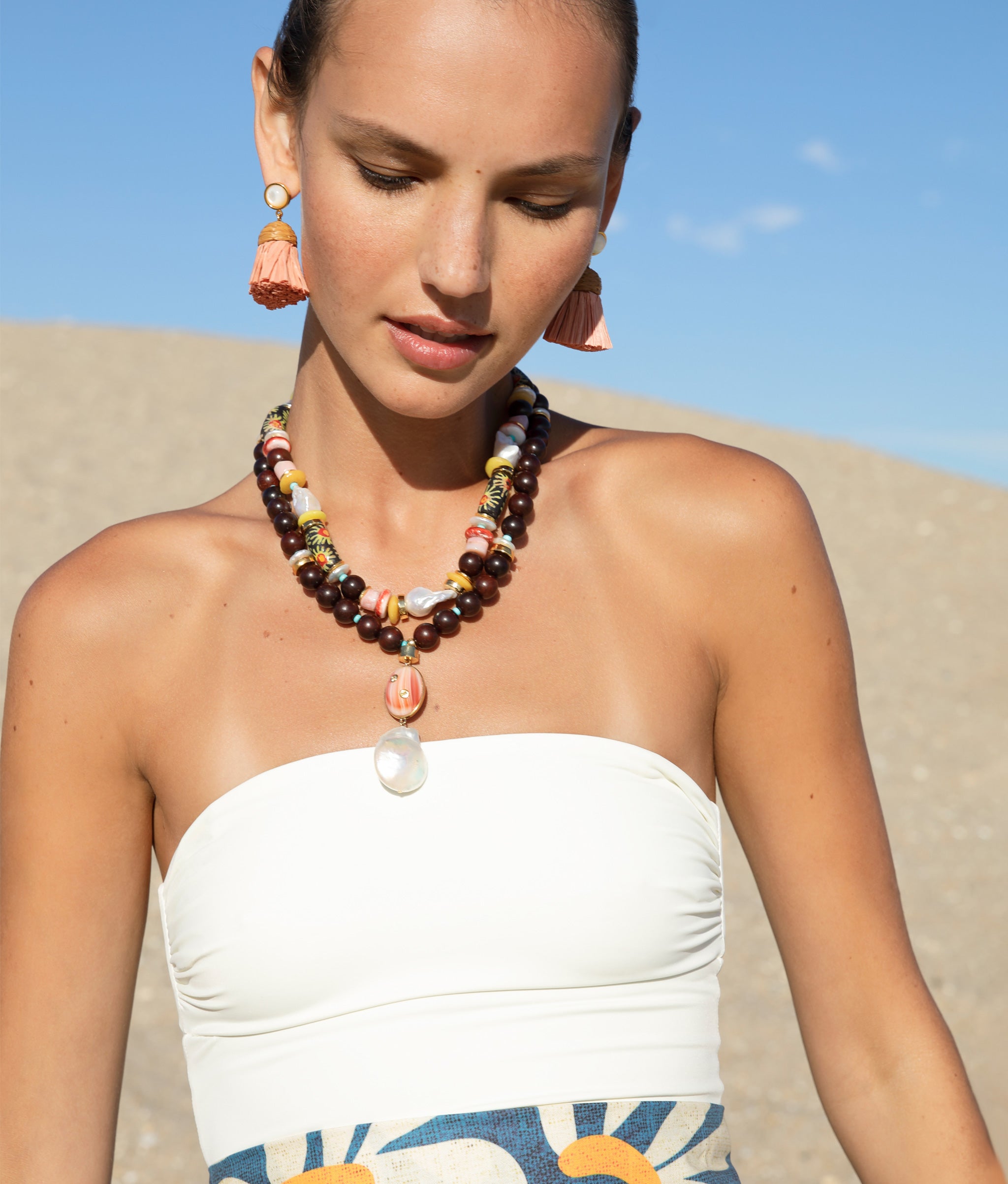 Model at the beach wearing the Raffia Earrings in Rose with the Gaia Necklace and the Souvenir Necklace in Tropic.