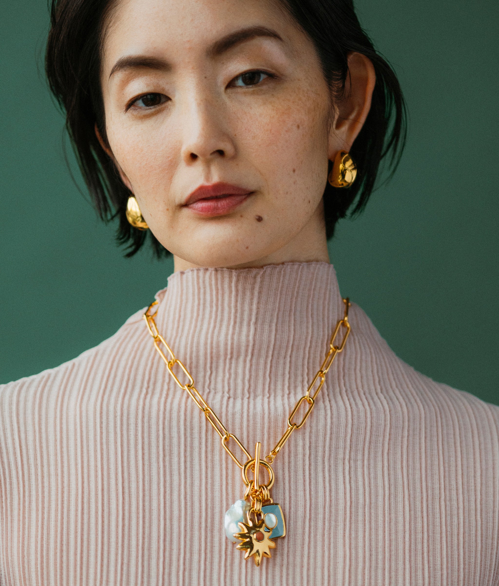 Model on green backdrop wears pink pleated top with Helios Charm Necklace and Mini Arp Earrings