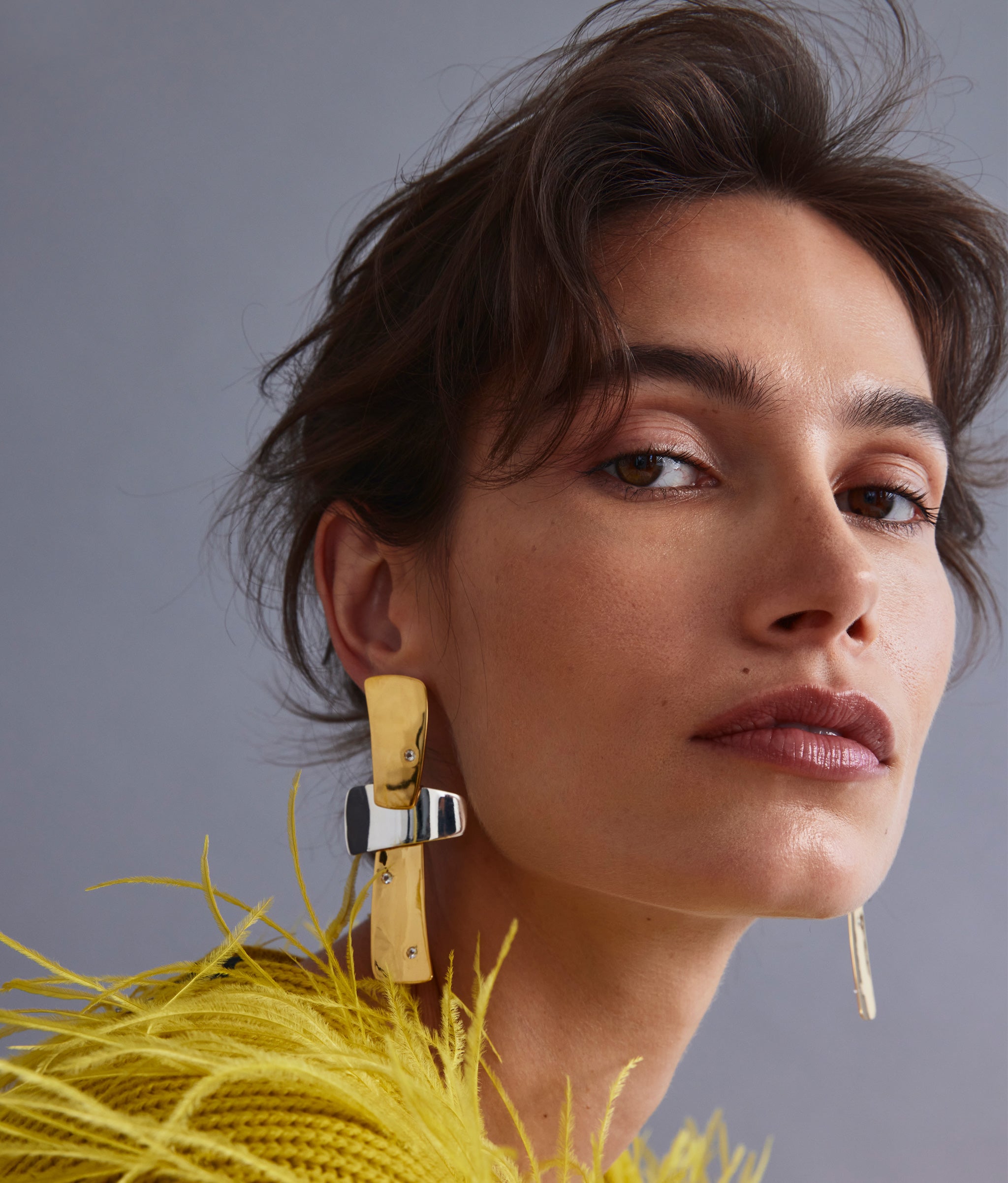 Model on grey backdrop wears feathery yellow top with Ernesto Earrings in Mixed Metal.