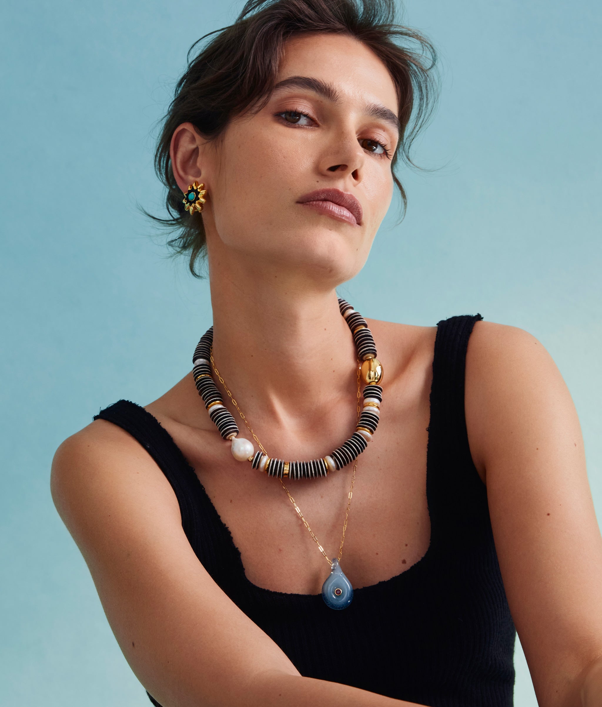 Model on blue backdrop wears black tank with Helios Stud Earrings, Prairie Necklace in Zebra and Muse Pendant in Rain.