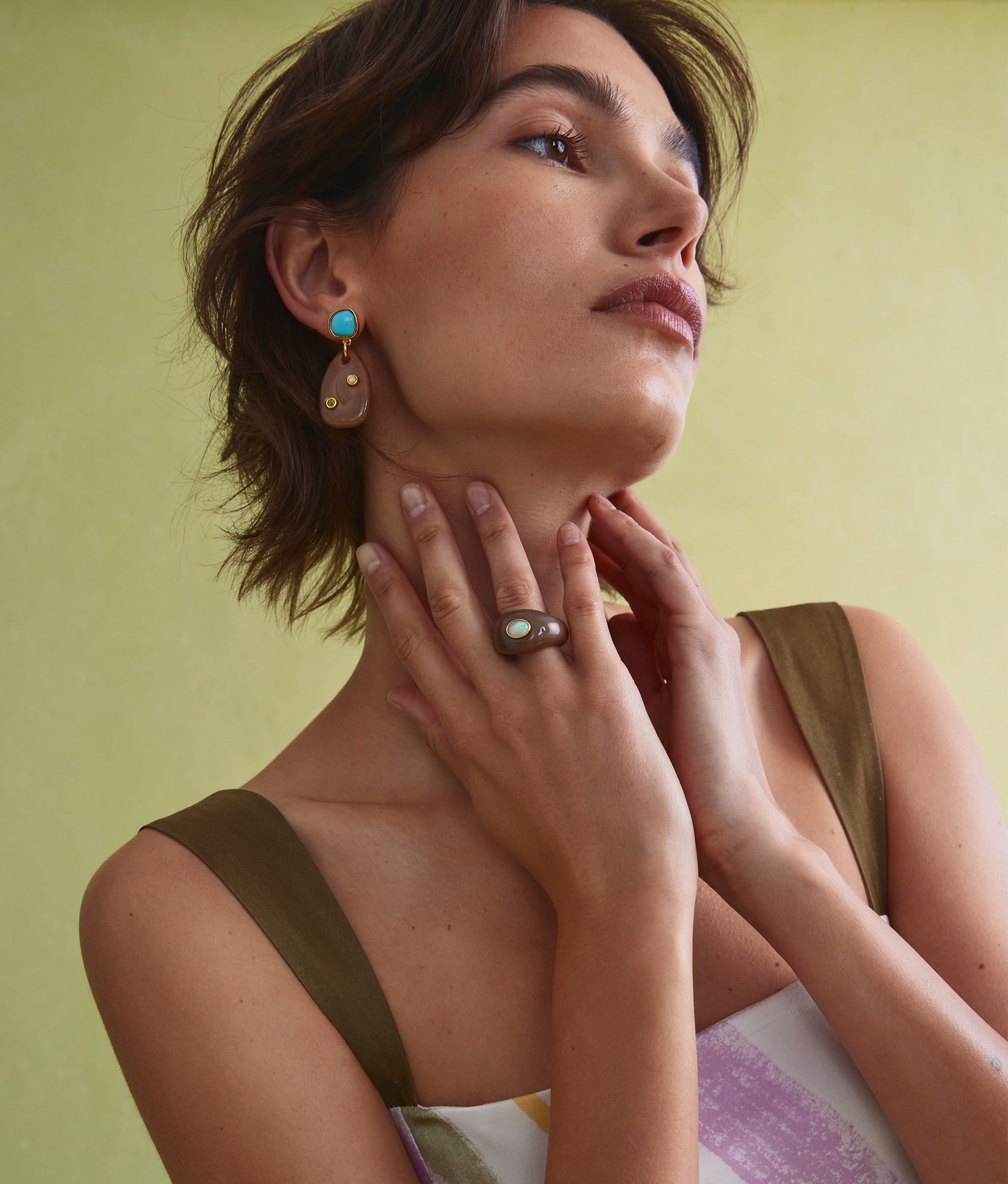 Model on green backdrop with hands on neck wears Monument Ring in Sandstone and Sandstone Earrings.