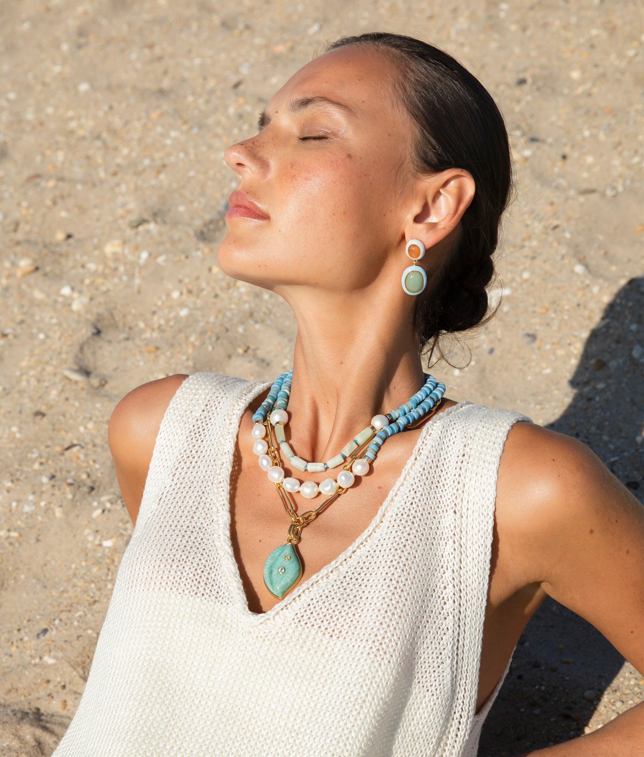 Model on the beach wearing Papaya Earrings paired with blue-toned, pearl, and gold necklaces.