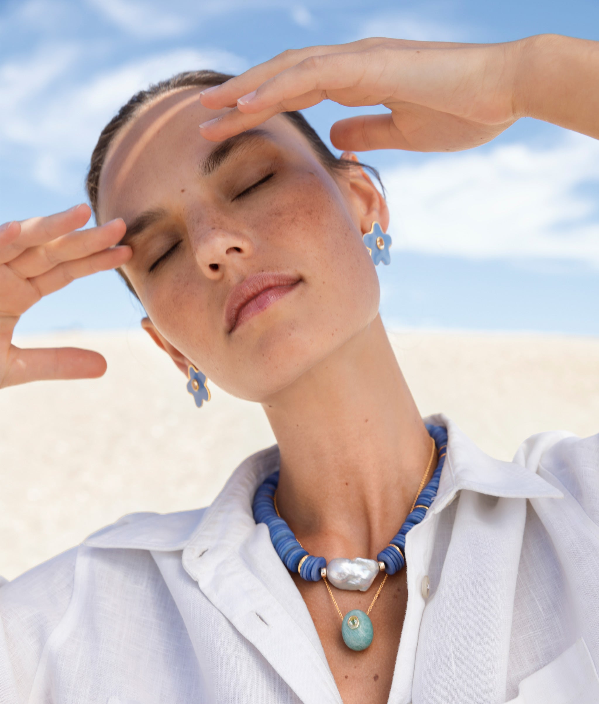 Model at the beach wearing Bilbao Collar in Marine paired with gold-chain necklace with blue stone and blue earrings.