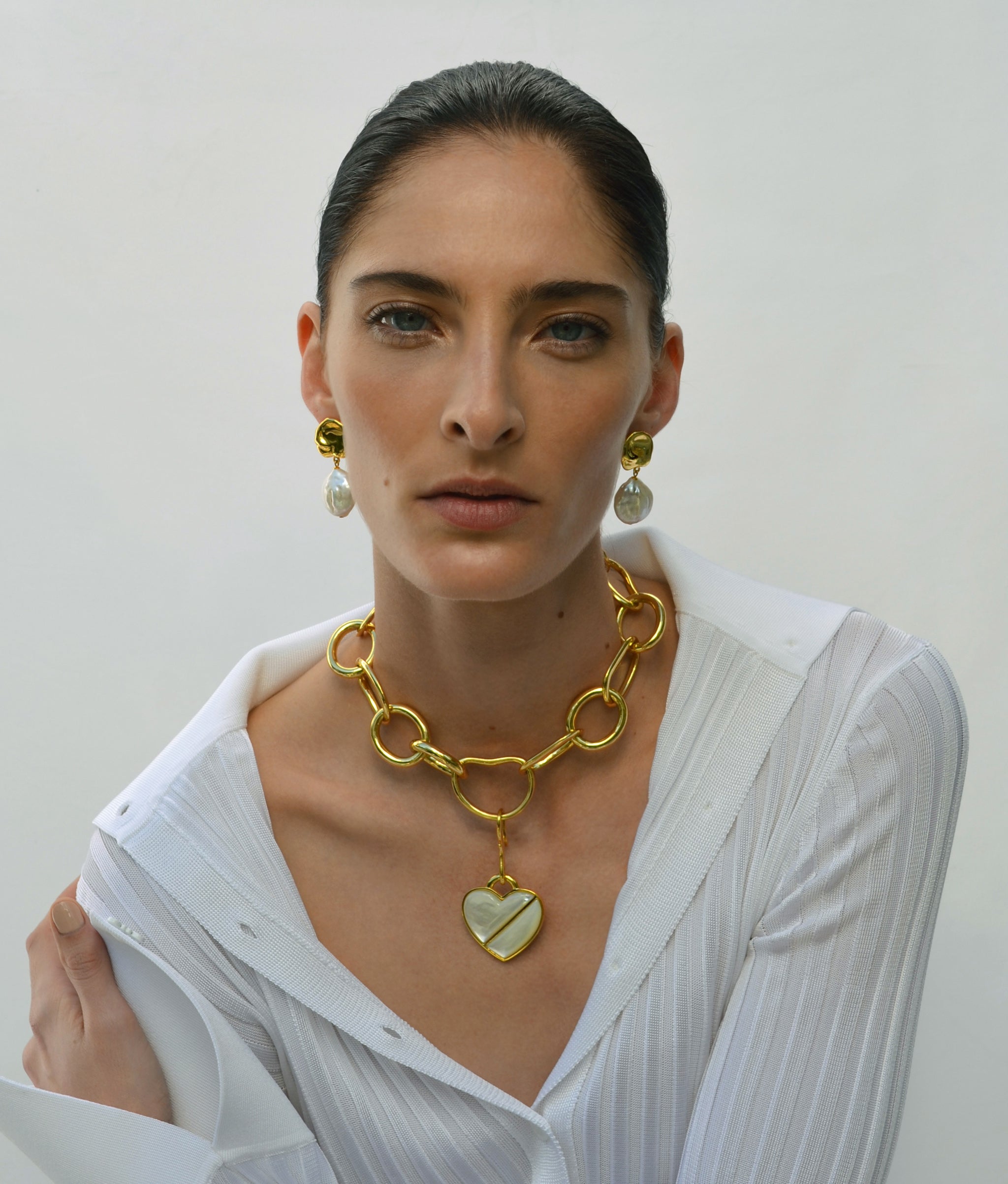 Model on grey backdrop wears white shirt with Porto Chain, Heart Pendant in Daydream, and Coin Reflection Earrings.