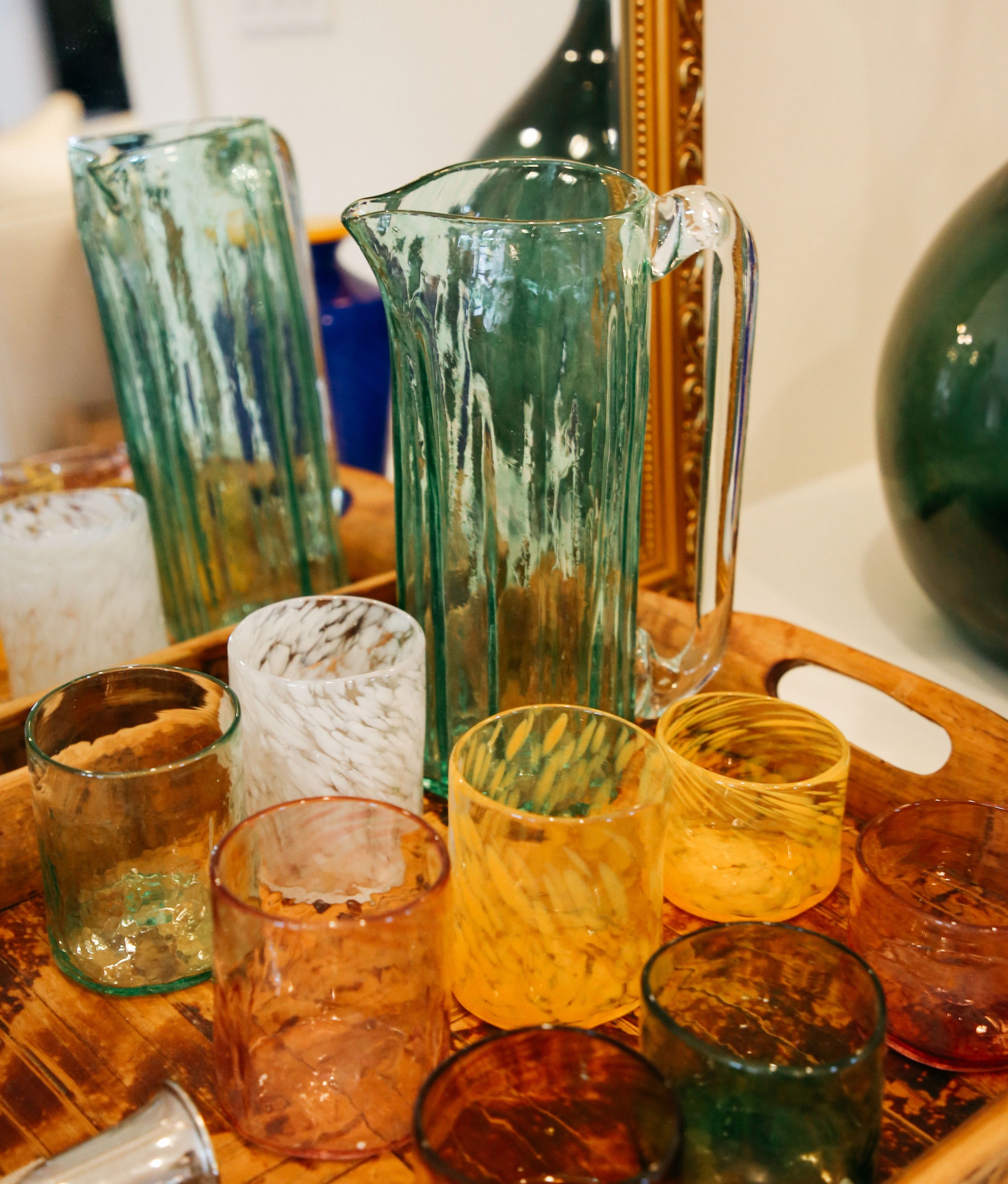 Wooden bar tray holds assorted colorful handblown glasses and pitcher.