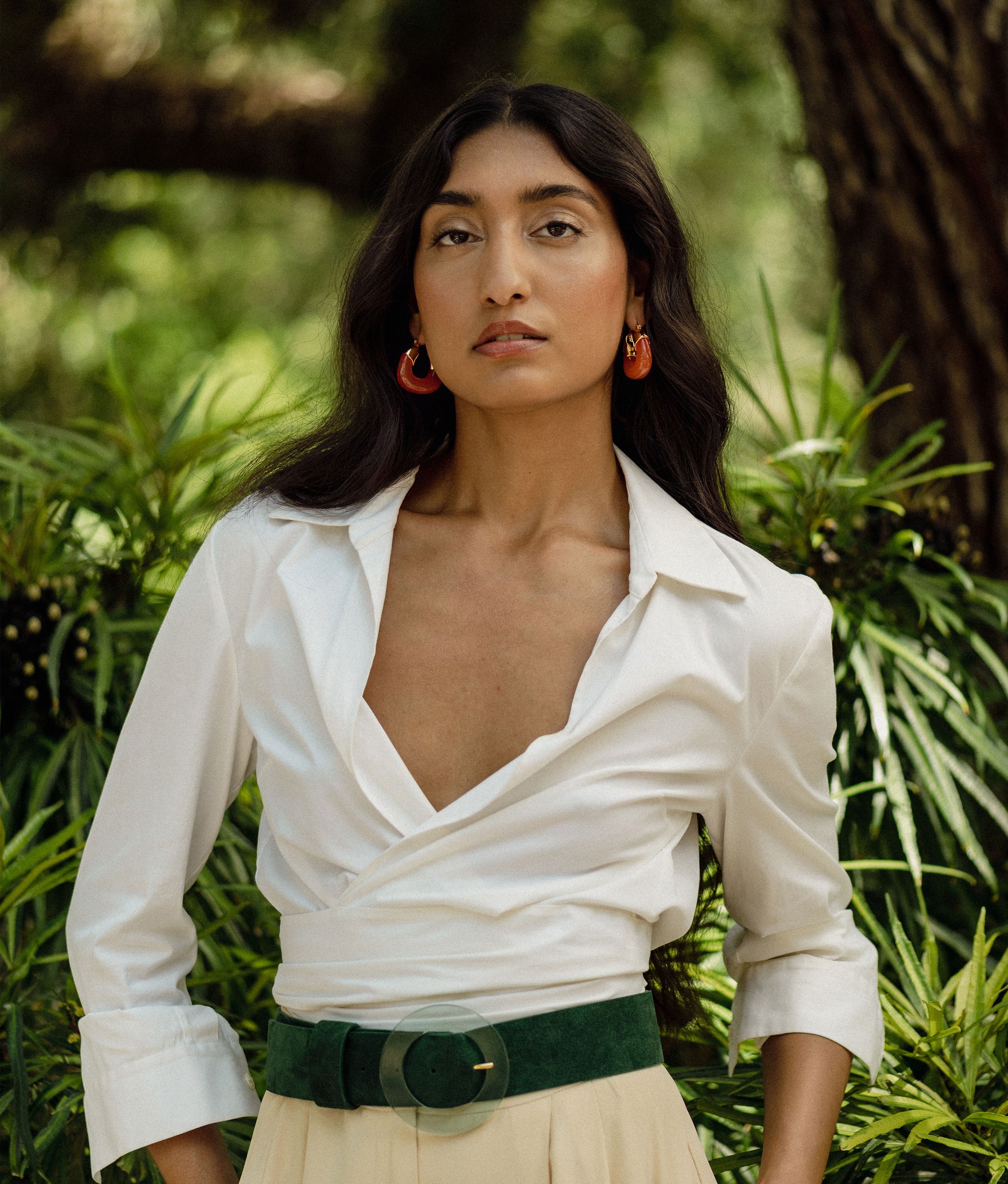 Model poses against green bushes in white top with Louise Belt in Emerald.