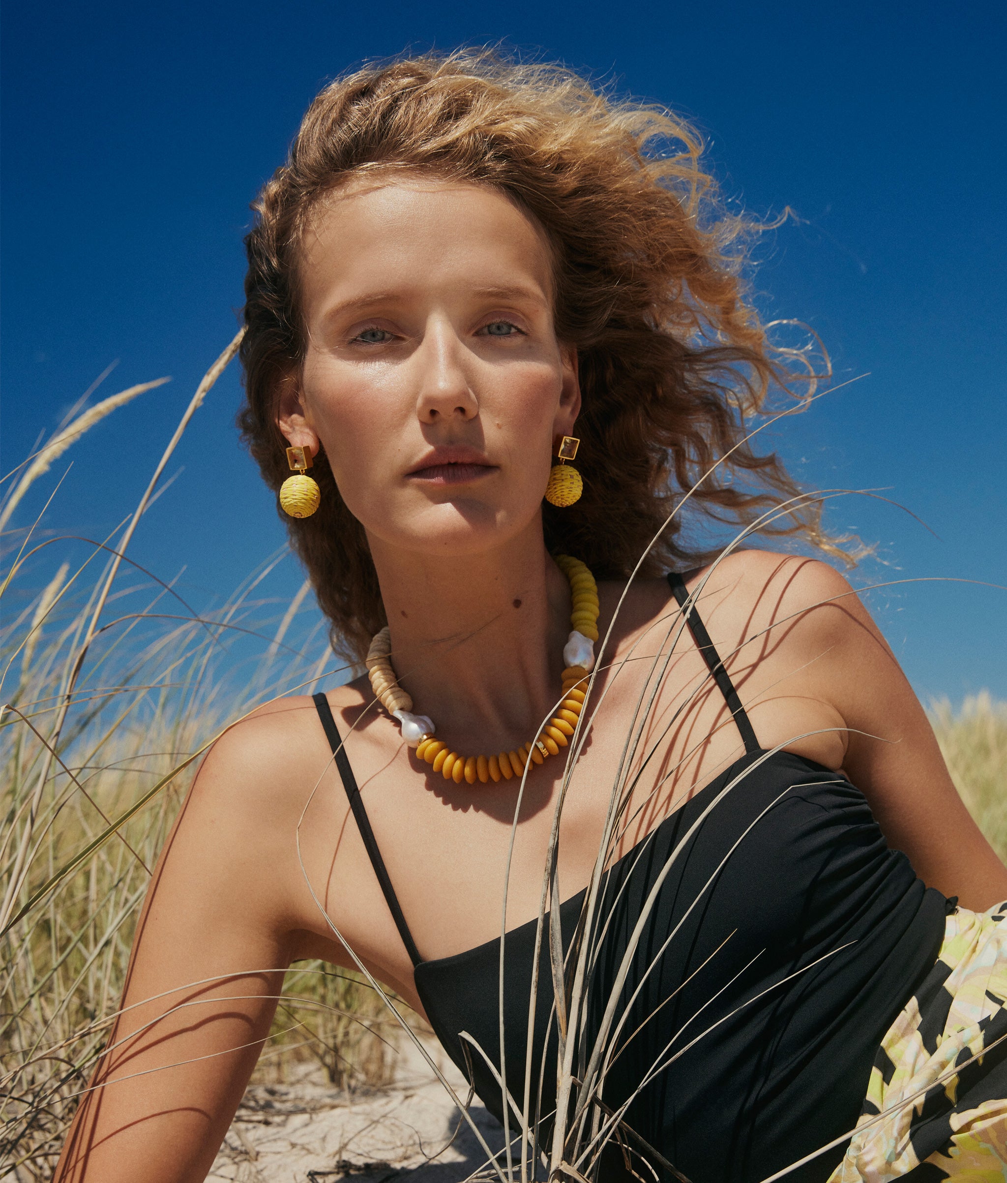 Model on beach dunes wears black swimsuit with Paradiso Earrings in Canary and Tavira Necklace in Sand.